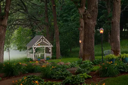 A white gazebo sits in a foggy, wooded yard with mature trees, garden beds, and glowing lamp posts.