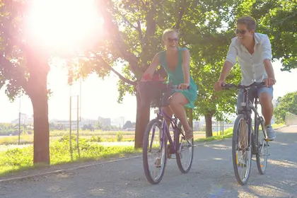 A man and woman ride bicycles on a sunlit path lined with trees.