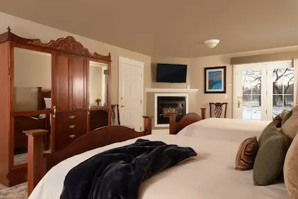 A hotel room with two beds, a wooden armoire, a fireplace with a mounted TV, and a door to the outdoors.