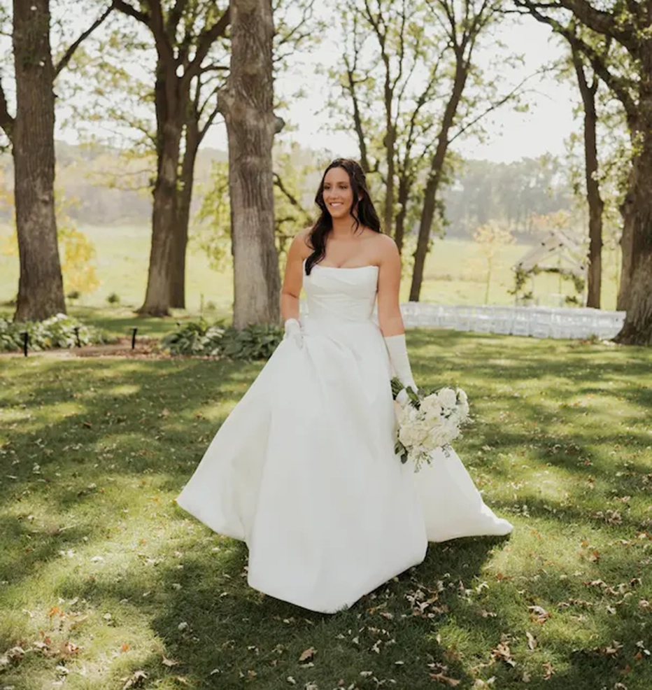 A bride in a strapless white gown and gloves walks across a grassy, tree-filled park, holding a white bouquet.