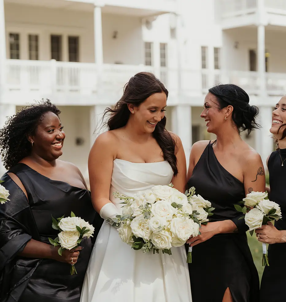 A bride in a white wedding gown holding flowers stands with bridesmaids in black dresses in front of a white building.