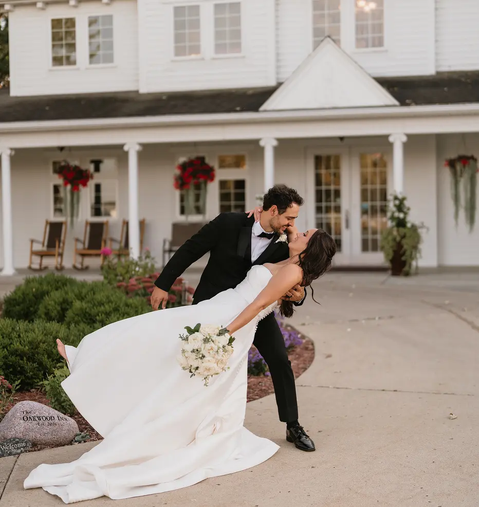 A groom in a tuxedo dips his bride in a white wedding gown in front of a white building.