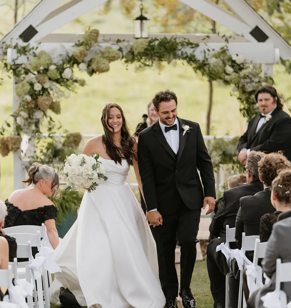A smiling bride in a white wedding dress and a groom in a black tuxedo walk hand-in-hand down an outdoor aisle.