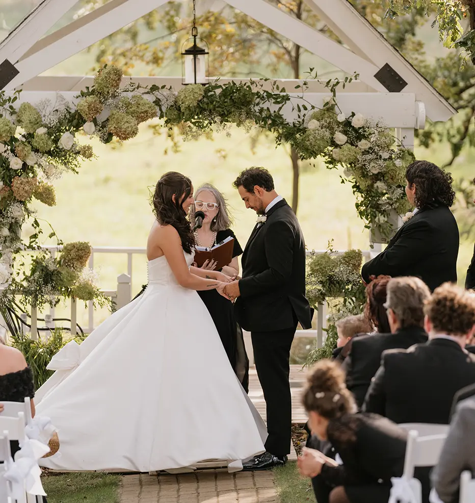 A bride and groom hold hands during their outdoor wedding ceremony under a white, flower-covered arch.