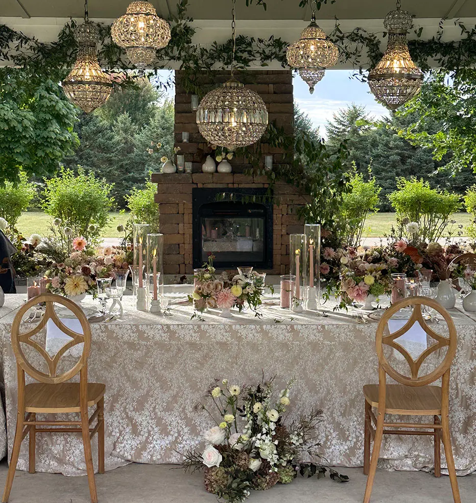 A beautifully decorated outdoor wedding table with lanterns hanging above a fireplace and garden backdrop.