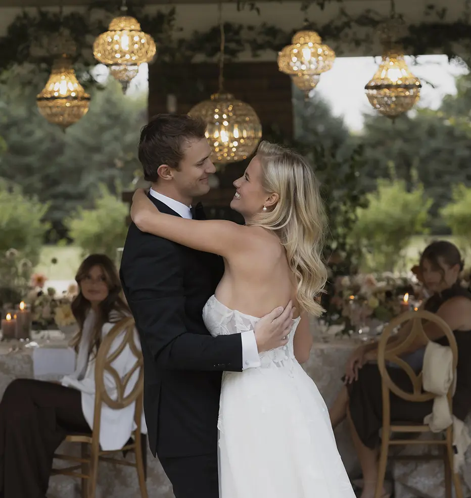 A bride and groom dancing together at a wedding reception, with guests seated at tables in the background.