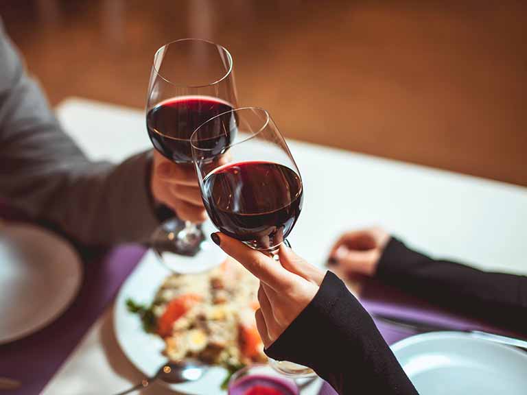Two people clink glasses of red wine over a meal at a dining table.