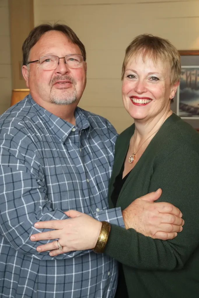 A middle-aged man in a blue plaid shirt and a woman in a green cardigan embrace and smile for a photo.