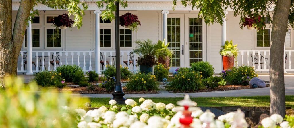 A white building with a porch, plants, and white flowers in the foreground.