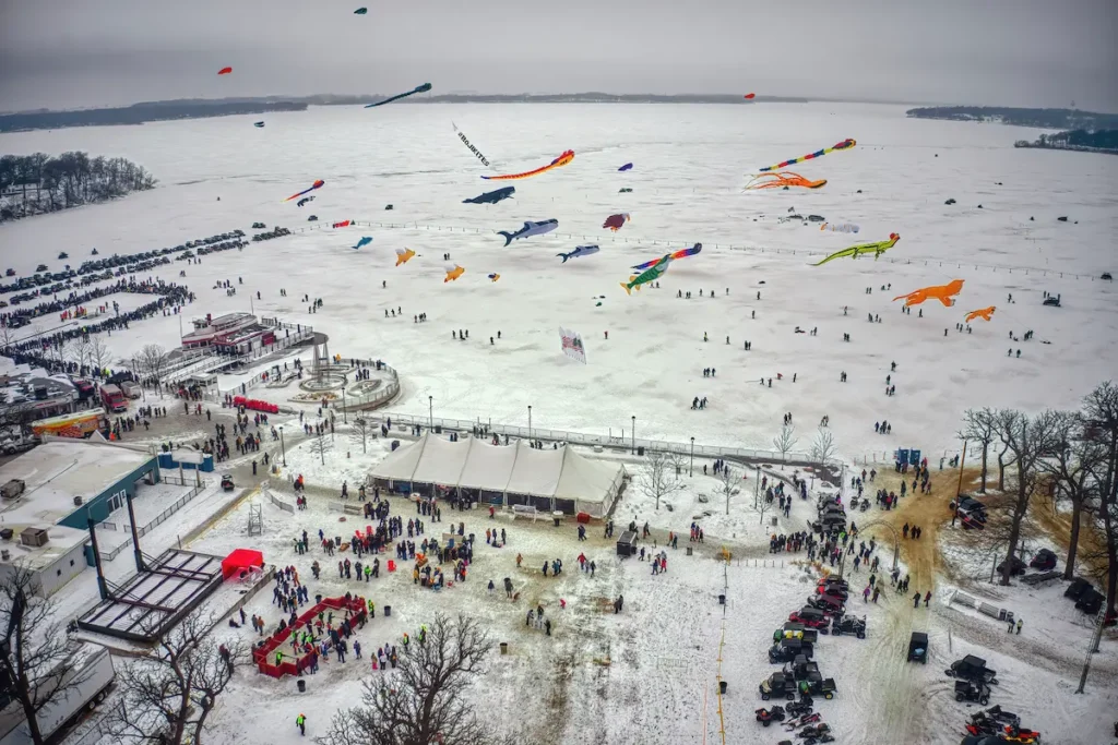 Aerial view of a snowy winter festival on a frozen lake with many colorful kites flying above a crowd of people.