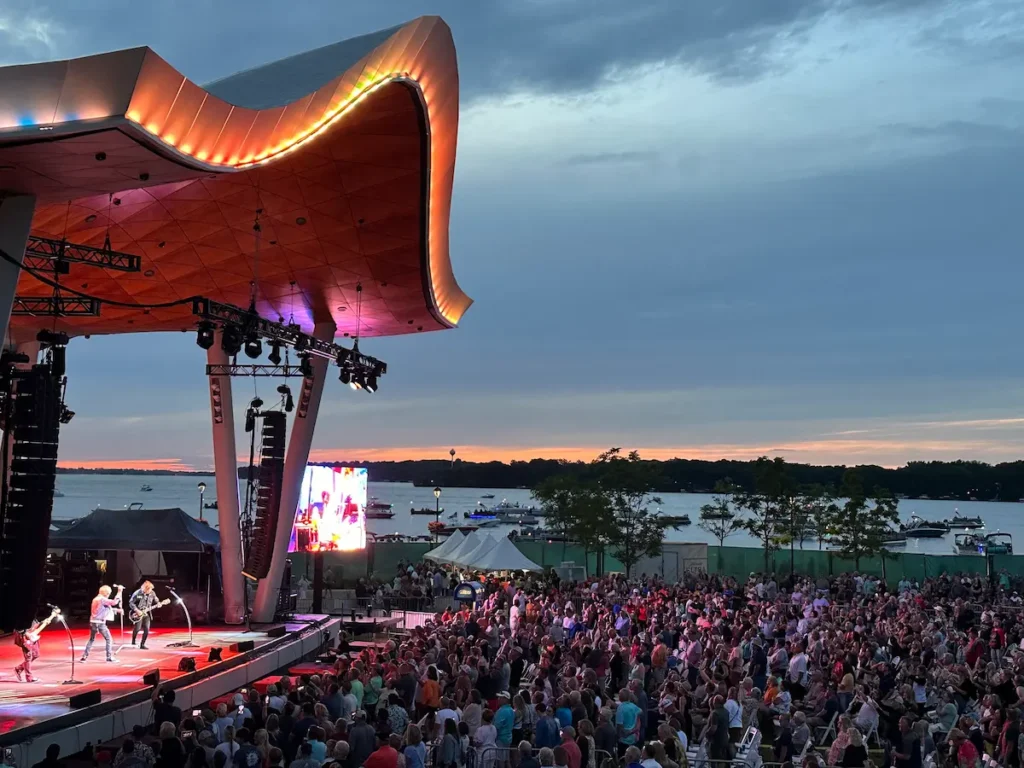A band performs on an outdoor stage at sunset before a large crowd gathered by a waterfront.