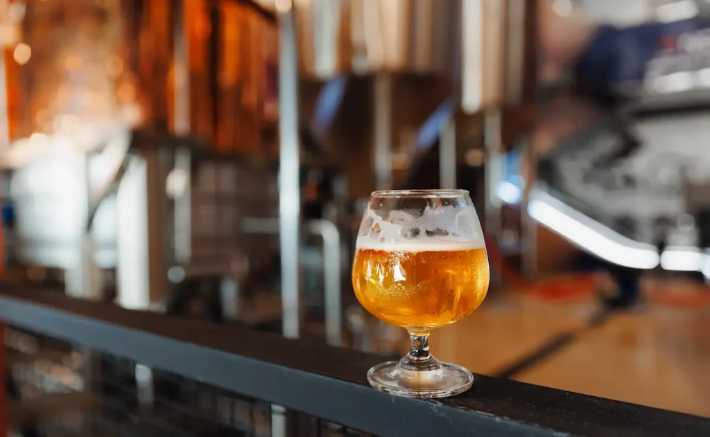 A glass of beer sits on a dark railing in front of a blurred brewery with large metal tanks.