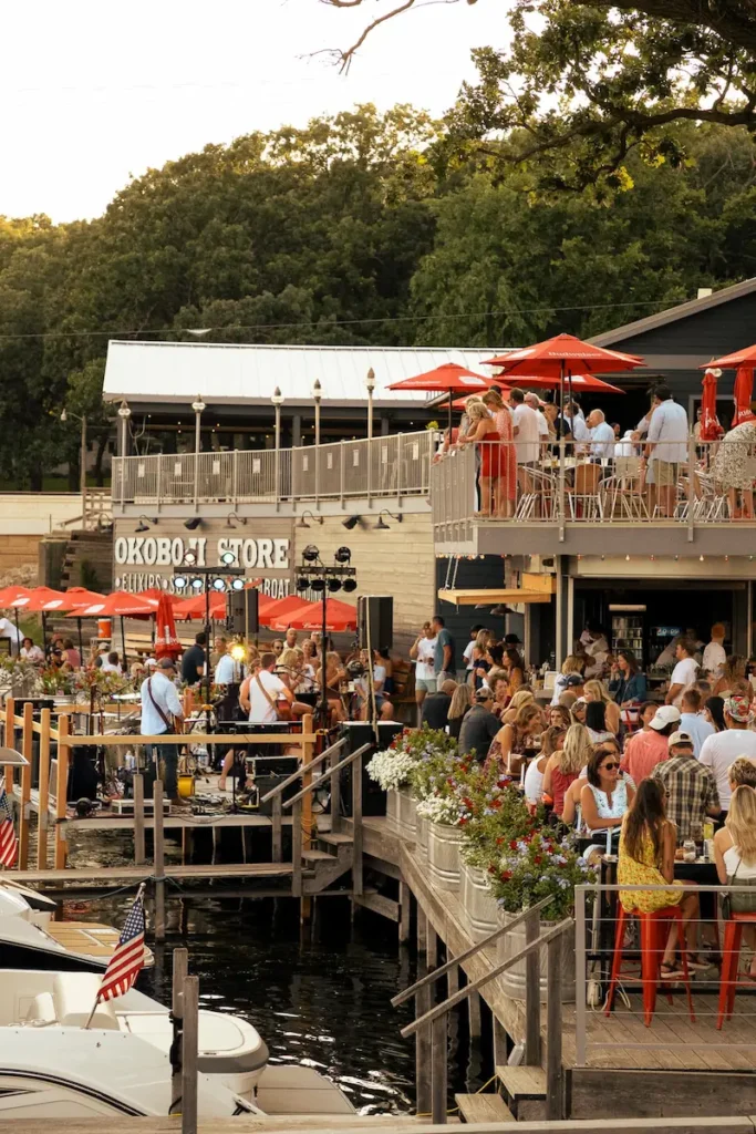 A crowd enjoys food and music at a waterfront restaurant with a deck, red umbrellas, and docked boats.