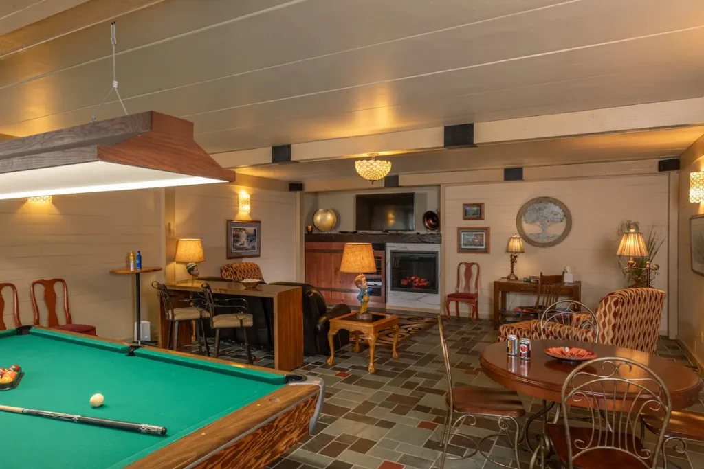 A spacious, rustic basement with a pool table, lounge area, fireplace, dining table, and stone-tiled floor.