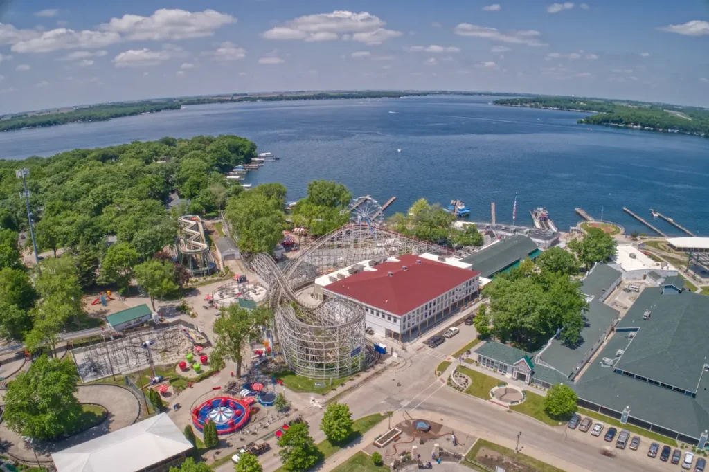 Aerial view of an amusement park with a wooden roller coaster next to a large lake under a clear blue sky.