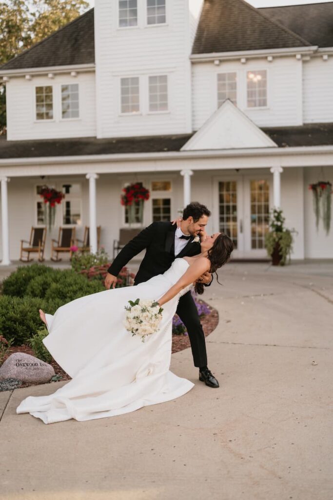 A groom in a tuxedo dips his bride in a white gown in front of a large, white estate house.